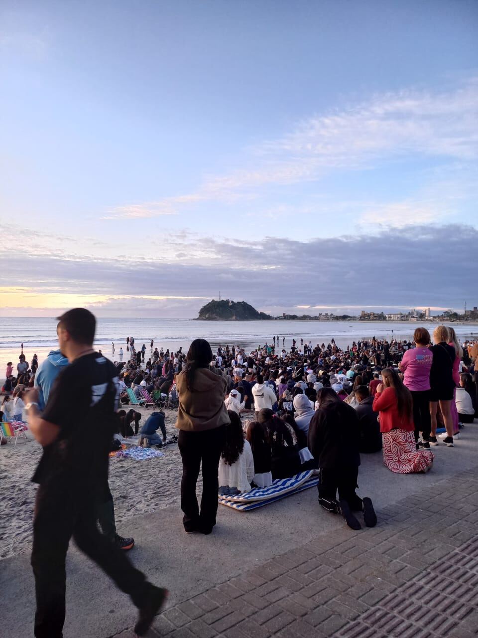 Pessoas reunidas no calçadão observando a oração coletiva na praia